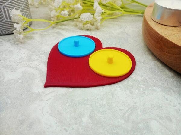 A romantic heart shaped toothbrush holder suitable for Philips toothbrushes. The image shows the red heart of the holder base with two bright circular holders of yellow and blue mounted on top. The two person heart shaped electric toothbrush holder is a modern, playful romantic gift for couples moving in together or as a fun romantic 'his and her' gift.