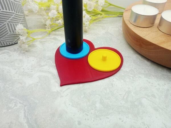 A romantic heart shaped toothbrush holder suitable for Philips toothbrushes. The image shows the red heart of the holder base with two bright circular holders of yellow and blue mounted on top. The two person heart shaped electric toothbrush holder is a modern, playful romantic gift for couples moving in together or as a fun romantic 'his and her' gift.