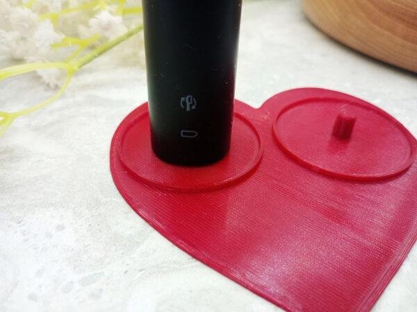 A side view of the two person Philips electric toothbrush holder. The classic, romantic heart shape holder 3d printed in a deep red, contrasting against the dark marble of the background. The small lip surrounding both holding posts can be seen. The design provides a snug fit with electric toothbrushes without the change of them getting stuck.