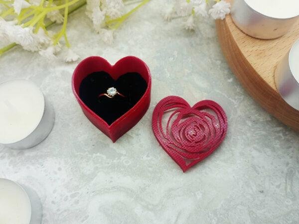 The size of the gift box is shown in comparison to the ring to see the scale (ring not included). The delicate pattering on the top of the link can be seen in the light gently reflecting off the lid. The curves of the heart shaped base can be seen against the marble tile.