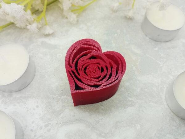 The romantic red, ring gift box is located on a marble back drop surrounded by tealights and flowers for perspective. The intricate and modern shaping of the box lid is displayed against the deep black of the ring insert behind. The links of the heart shaped box and rose filigree in red are clearly reminiscent of valentine's day and other romantic events.