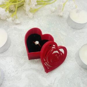 A ring presentation gift box shaped like a heart with a rose filigree outline cut into the lid. Where the filigree leaves gaps in the lid you can glimpse the rich black of the ring cushion. The gentle shape of the rose is created by very fine 3d printing with some of the lines measuring only just over 1mm in width. It is displayed next to some standard size tealights to give a sense of scale.