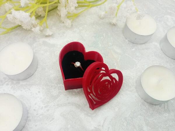 A ring presentation gift box shaped like a heart with a rose filigree outline cut into the lid. Where the filigree leaves gaps in the lid you can glimpse the rich black of the ring cushion. The gentle shape of the rose is created by very fine 3d printing with some of the lines measuring only just over 1mm in width. It is displayed next to some standard size tealights to give a sense of scale.