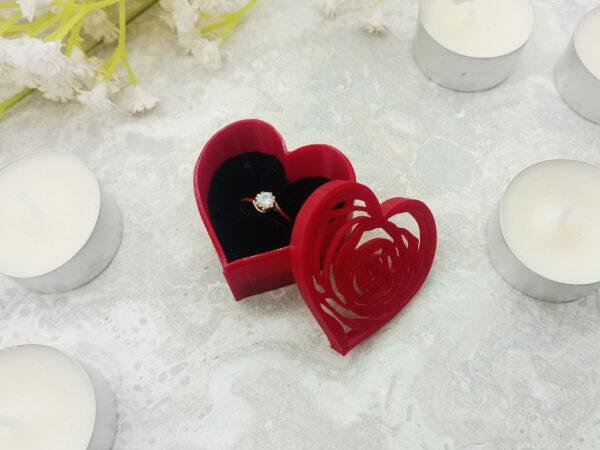 The red heart shaped ring gift box is surrounded with tealights to give a sense of scale. The lid for the box is leaning against the box so you can see the depth of the lid and the box. The red of the box is contrasted by the black of the ring display foam that holds the ring upright. The ring fits nicely into the box, leaving enough room not to be cramped but enough room to show the ring to its best advantage. The ring is not included.